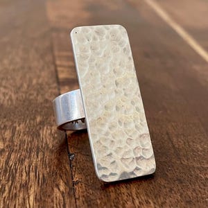 May include: A silver ring featuring a rectangular hammered metal face. The ring band is smooth silver. The textured face has a light, neutral colour, and the ring is set against a wooden surface.