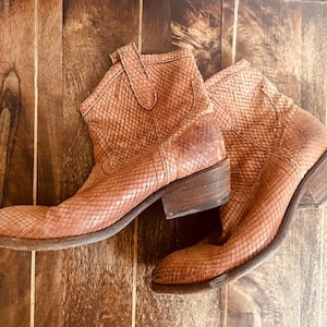 Könnte beinhalten: Ein Paar braune Leder-Ankle-Boots mit Schlangenmuster. Die Stiefel haben eine spitze Zehenpartie, einen niedrigen Absatz und eine Zuglasche oben. Die Stiefel stehen vor einem hölzernen Hintergrund.