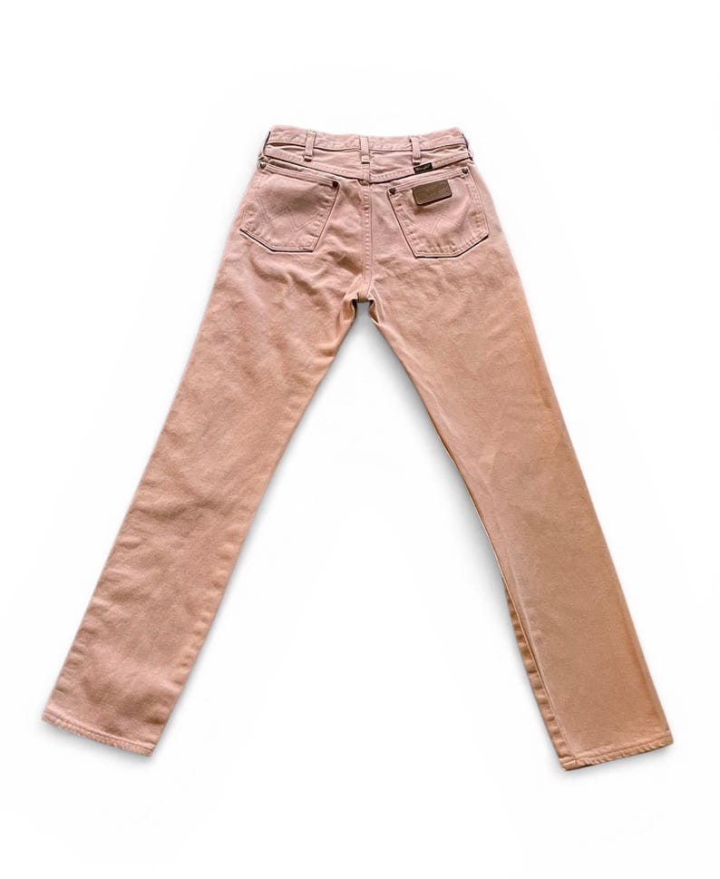 May include: A pair of light pink denim jeans featuring a high-rise waist, a classic five-pocket design, and a brown leather patch on the back right pocket. The jeans have a straight-leg cut with visible stitching and metal rivets throughout.