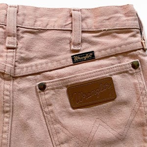 May include: A pair of light pink denim jeans featuring a high-waisted design with belt loops, a black Wrangler brand tag, and a brown leather patch on the back pocket. The pocket is reinforced with metallic rivets and displays a stitched W pattern.