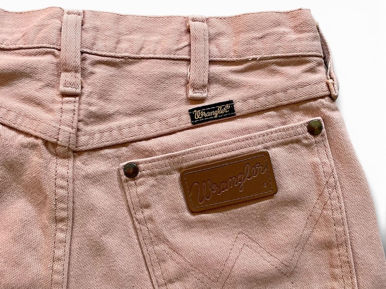 May include: A pair of light pink denim jeans featuring a high-waisted design with belt loops, a black Wrangler brand tag, and a brown leather patch on the back pocket. The pocket is reinforced with metallic rivets and displays a stitched W pattern.