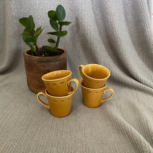 May include: Four yellow ceramic mugs stacked on top of each other. The mugs have a scalloped rim and a textured surface. The mugs are sitting on a gray textured surface.