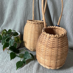 May include: Two woven baskets with arched handles. The baskets are made of light brown wicker and have a rounded shape. The handles are attached to the top of the baskets. A small potted plant with green leaves is to the left of the baskets.