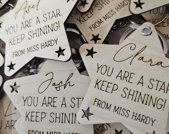 Puede incluir: Primer plano de varios llaveros de madera en forma de estrella con el texto "You are a star. Keep shining! From Miss Hardy" en cada llavero. Los llaveros están en una pila y los nombres Asel, Josh y Clara son visibles.