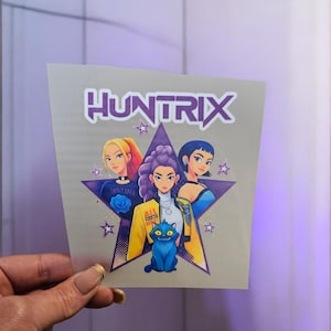 May include: A sticker with the word "HUNTRIX" above a star graphic. The star contains three stylized female figures and a blue cat. The figures have different hair colours and wear various outfits. The background is a light purple.