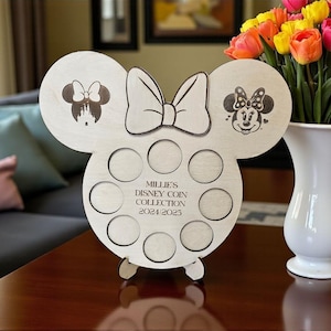 Puede incluir: Expositor de monedas de madera con forma de cabeza de Mickey Mouse, con imágenes grabadas de Minnie Mouse y un lazo. Tiene ranuras circulares para monedas y el texto "Millie's Disney Coin Collection 2021/2023".