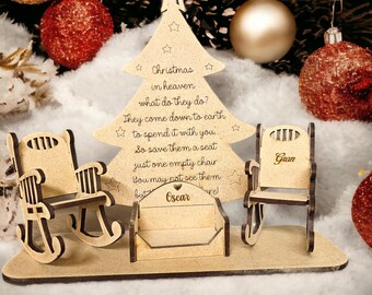 Puede incluir: Un árbol de Navidad de madera con una estrella en la parte superior y texto grabado, flanqueado por mecedoras en miniatura y un banco. El árbol está rodeado de adornos navideños dorados y rojos, creando una escena festiva.