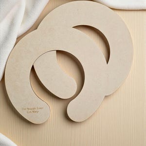 May include: Two unfinished wooden craft shapes, each with a curved design and a rounded end. The text "The Snappy Laser Cut Shop" is visible on one piece. The shapes are light brown and rest on a light wood surface.