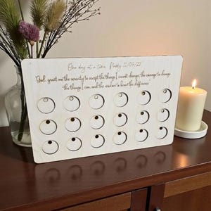 May include: A wooden plaque with a series of circular cutouts and engraved text. The text reads "One day at a time. Paddy 11/09/22" and a prayer. A lit candle and a vase of flowers are in the background.
