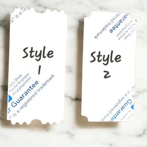 May include: Two white rectangular tags with rounded corners and a scalloped edge. The text "Style 1" and "Style 2" are written in black marker. The tags have the text "Guarantee" and "is a registered trademark".