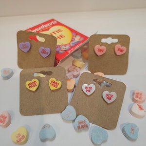 May include: A set of six pairs of heart-shaped earrings with different sayings on them. The earrings are displayed on brown card stock with a box of Sweethearts candies in the background. The sayings on the earrings are "Go Away", "Hard Pass", "Bite Me", and "EW".
