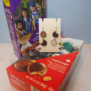May include: A pair of wooden earrings shaped like Girl Scout Tagalongs cookies. The earrings are on a wooden display stand. The earrings are brown and black with a white swirl design. The earrings are a fun and unique way to show your love for Girl Scout cookies.