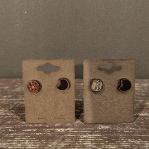 May include: Two pairs of wooden stud earrings on brown cardboard earring cards. One pair features a moon and a circular design, the other pair has a crescent moon and a circular design. The text "Chaos Creates" is written on the cards.