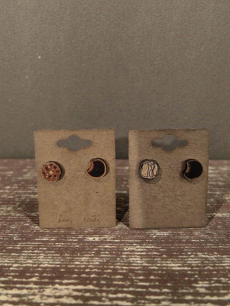 May include: Two pairs of wooden stud earrings on brown cardboard earring cards. One pair features a moon and a circular design, the other pair has a crescent moon and a circular design. The text "Chaos Creates" is written on the cards.
