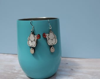 Pode incluir: Um par de brincos pendentes com uma galinha branca com crista vermelha e um pequeno amuleto de ovo de madeira. Os brincos são prateados e têm um design simples.
