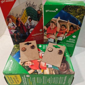 May include: Girl Scout cookie boxes, including Tagalongs and Thin Mints, with two pairs of wooden earrings. The earrings are shaped like cookies. The boxes are red and green with images of girls.