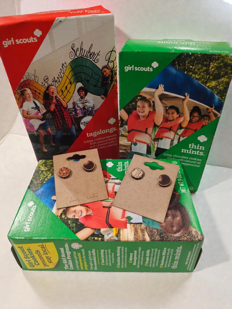 May include: Girl Scout cookie boxes, including Tagalongs and Thin Mints, with two pairs of wooden earrings. The earrings are shaped like cookies. The boxes are red and green with images of girls.