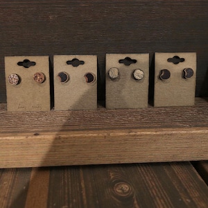 May include: Four pairs of wooden stud earrings displayed on brown cardboard backing. The earrings feature various designs, including textured patterns, crescent moons, and wave-like lines. The earrings are on a wooden surface.