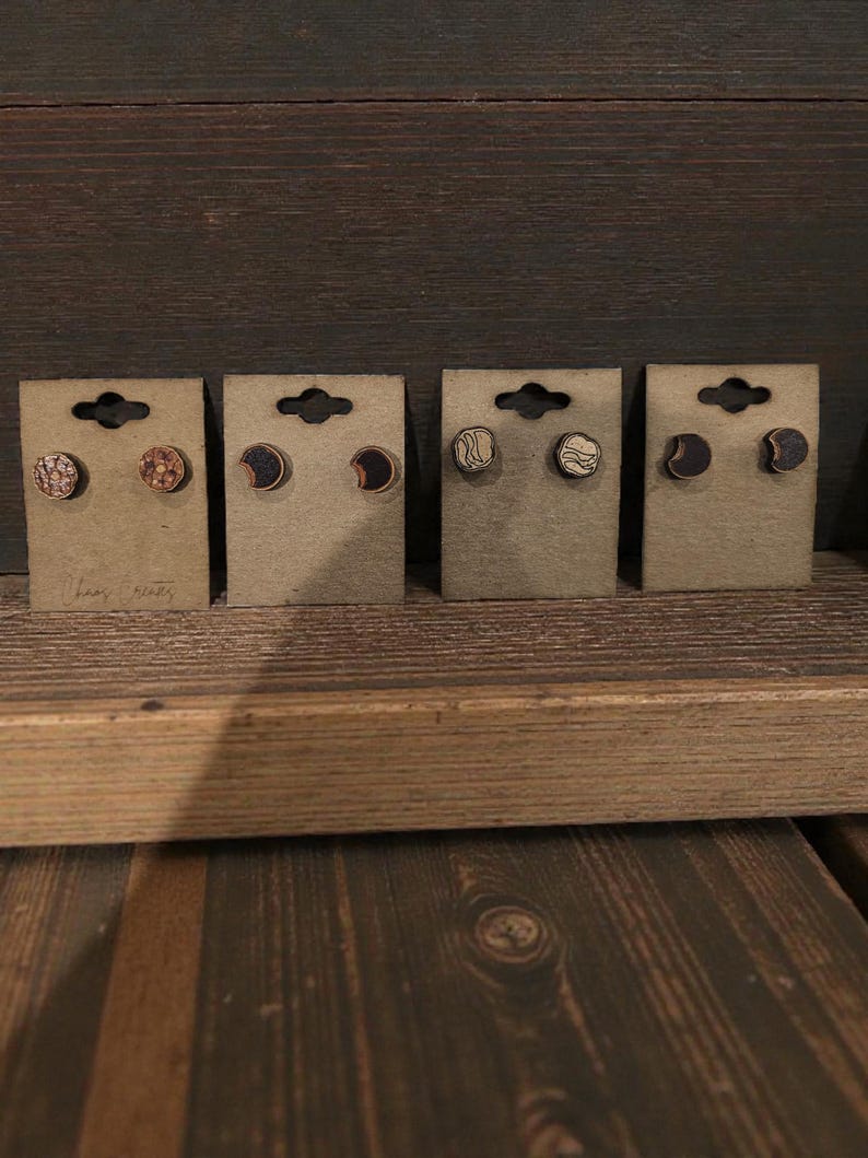 May include: Four pairs of wooden stud earrings displayed on brown cardboard backing. The earrings feature various designs, including textured patterns, crescent moons, and wave-like lines. The earrings are on a wooden surface.
