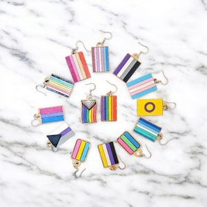 May include: A collection of rectangular earrings with various LGBTQ+ pride flag designs. The earrings feature gold-colored hooks and are arranged on a white marble surface. The flags display a range of colors, including rainbow stripes, pink, blue, purple, and yellow.