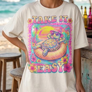 May include: Cream-colored t-shirt with a colorful graphic of a turtle relaxing on an inflatable ring at the beach. The graphic includes the text "TAKE IT EASY" in pink and yellow. Palm trees, flowers, and a sunset are also featured.