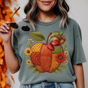 May include: A sage green t-shirt featuring a colorful apple design. The apple is composed of various patterned sections in orange, yellow, and green, with red leaves, acorns, and yellow flowers. The shirt is a comfortable, casual style.