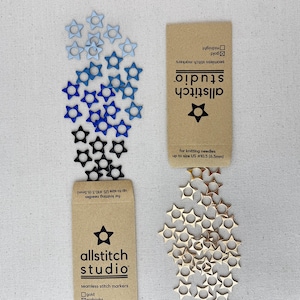 May include: Star-shaped stitch markers in shades of blue, black, and gold are displayed. The markers are scattered around two product cards. The cards read "allstitch studio" and "seamless stitch markers" and are for knitting needles up to size US #10.5 (6.5mm).