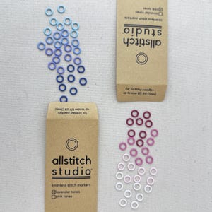 May include: Two packages of seamless stitch markers for knitting needles up to size US #8 (5mm). The packages are brown and feature the text "allstitch studio" and a circle logo. The markers are in shades of blue, purple, pink, and white.