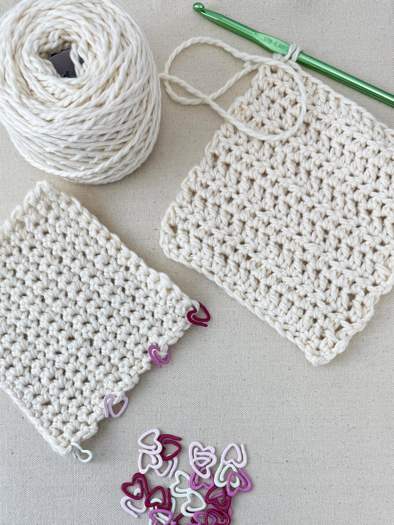 May include: Two crocheted squares made with cream colored yarn, a ball of cream colored yarn, a green crochet hook, and a pile of pink and red heart shaped stitch markers.