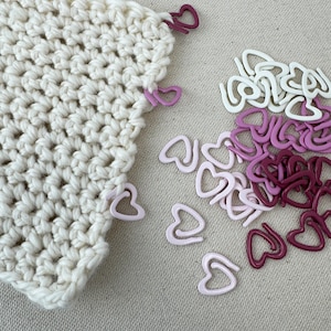 May include: A close-up of a white crocheted fabric with pink and burgundy heart-shaped stitch markers. The markers are scattered on a beige surface.