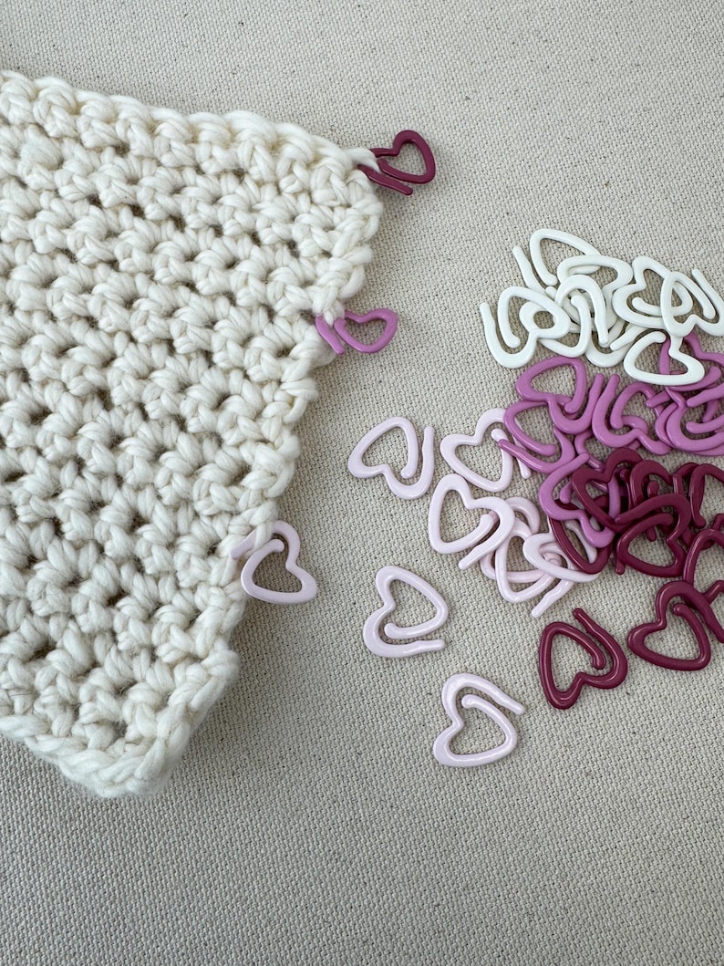 May include: A close-up of a white crocheted fabric with pink and burgundy heart-shaped stitch markers. The markers are scattered on a beige surface.