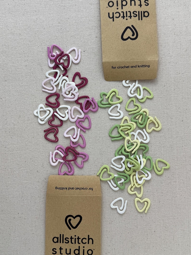 May include: Assortment of colorful heart-shaped stitch markers in pink, green, and white. The markers are arranged in a pile on a beige surface. The image also includes two brown paper envelopes with the text "allstitch studio" and "for crochet and knitting".