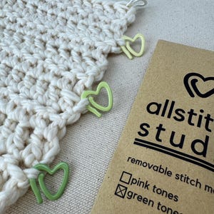 May include: A close-up of a crocheted project with green heart-shaped stitch markers. The project is on a beige surface. A brown card with the text "allstit studio" and "removable stitch markers" is in the lower right corner. The card has checkboxes for "pink tones" and "green tones", with the green tones box checked.