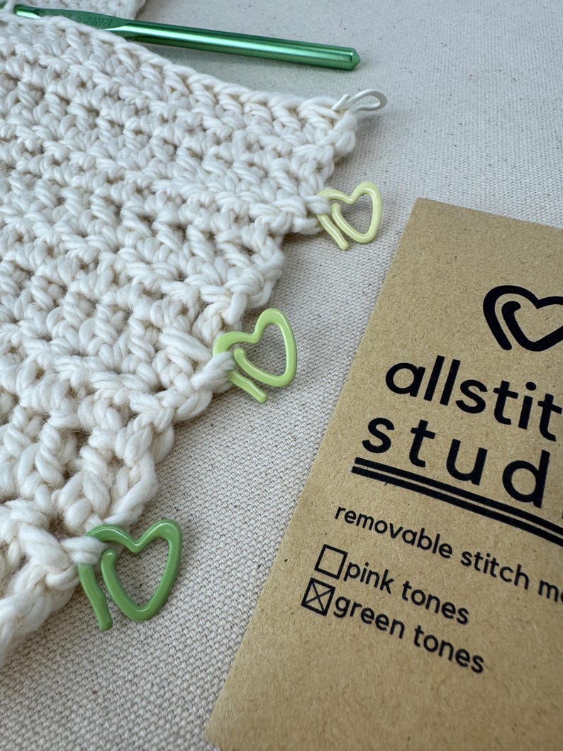 May include: A close-up of a crocheted project with green heart-shaped stitch markers. The project is on a beige surface. A brown card with the text "allstit studio" and "removable stitch markers" is in the lower right corner. The card has checkboxes for "pink tones" and "green tones", with the green tones box checked.