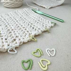 May include: White crochet fabric with green and yellow heart-shaped stitch markers. The fabric is laid out on a beige surface.
