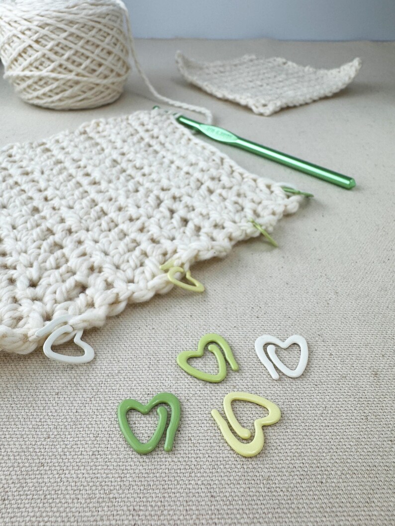 May include: White crochet fabric with green and yellow heart-shaped stitch markers. The fabric is laid out on a beige surface.