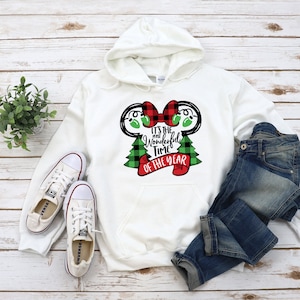 Puede incluir: Sudadera con capucha blanca con un diseño de cuadros rojos y verdes que presenta una cabeza de Mickey Mouse con un lazo rojo y el texto "It's the most Wonderful Time of the Year".