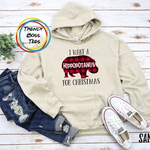 Puede incluir: Una sudadera con capucha beige con un gráfico de hipopótamo de cuadros rojos y negros y el texto "Quiero un hipopótamo para Navidad".