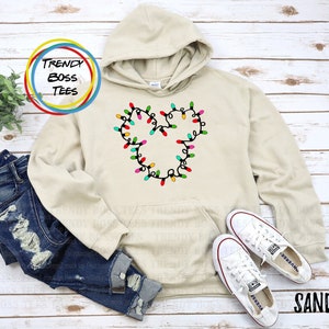 Puede incluir: Una sudadera con capucha de color arena con un diseño en forma de Mickey Mouse hecho de luces navideñas de colores. La sudadera tiene una capucha con cordón y un bolsillo canguro.