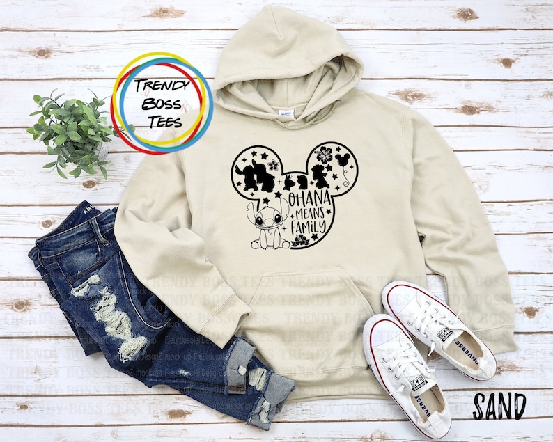 K&ouml;nnte beinhalten: Sandfarbener Hoodie mit einem schwarzen Mickey-Maus-Kopf-Design. Das Design beinhaltet Stitch, Lilo und andere Figuren aus dem Disney-Film Lilo & Stitch. Der Text "Ohana means family" befindet sich innerhalb des Mickey-Maus-Kopfes.