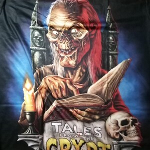 May include: A black t-shirt featuring a graphic of a skeletal figure reading a book titled "Tales from the Crypt". The figure is seated on a throne with a skull on the armrest. A lit candle is on the left side of the image.
