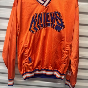 May include: An orange V-neck pullover jacket featuring the word KNICKS in large purple letters across the chest. The garment has blue and white striped trim on the collar, cuffs, and hem, with a small NBA logo and a Starter brand logo visible.