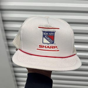 May include: White and mesh New York Rangers baseball cap with a red brim and the Sharp logo. The cap features the team's logo in red, white, and blue.
