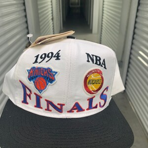 Könnte beinhalten: Weiße Baseballkappe mit schwarzem Schirm und grünem Unterbrim. Die Kappe hat ein blaues und weißes New York Knicks Logo, ein rot-weiß-gelbes Houston Rockets Logo und den Text "1994 NBA FINALS".