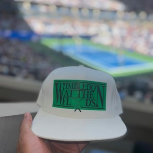 Op de afbeelding: Witte baseballcap met een groen rechthoekig ontwerp met de tekst "Wimbledon at the Floods". De cap heeft een gebogen klep en wordt omhoog gehouden tegen een vage achtergrond.