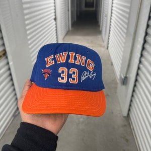 Puede incluir: Una gorra de béisbol azul y naranja con el texto "EWING 33" y una firma en tinta azul. La gorra tiene un logotipo de los New York Knicks en el lateral.