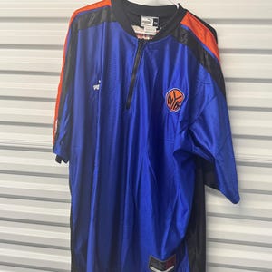 Puede incluir: Una camiseta de manga corta azul real con un diseño de bloques de color negro y naranja. Presenta una cremallera frontal parcial, un logotipo de Puma en el pecho y un emblema circular naranja y azul. Una etiqueta de la marca Puma es visible.