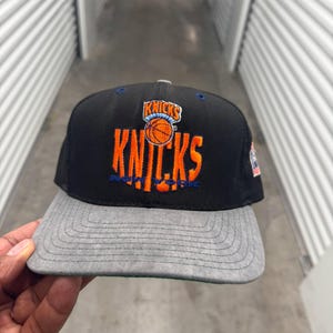 Puede incluir: Gorra de béisbol negra y gris de los New York Knicks. La gorra presenta el logotipo del equipo y la palabra "Knicks" en naranja y azul. La visera es gris y la corona es negra.