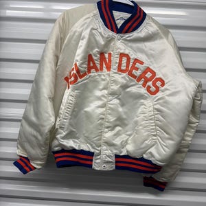 May include: A white satin baseball jacket with blue and red striped trim. The jacket has a snap closure and features the word "Islanders" in orange lettering on the front.