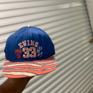 Puede incluir: Una gorra de béisbol azul y naranja con el texto "EWING 33" y el logotipo de los New York Knicks. La gorra tiene un patrón a rayas en la visera.